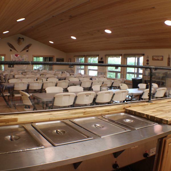Steam table serving line in the Camp Maranatha Dining Hall, with dining room seating and lake facing windows beyond.