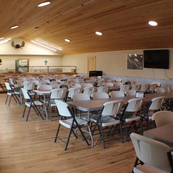 Dining hall interior with rows of tables and chairs, wall mounted TV, and serving counter at the far end.